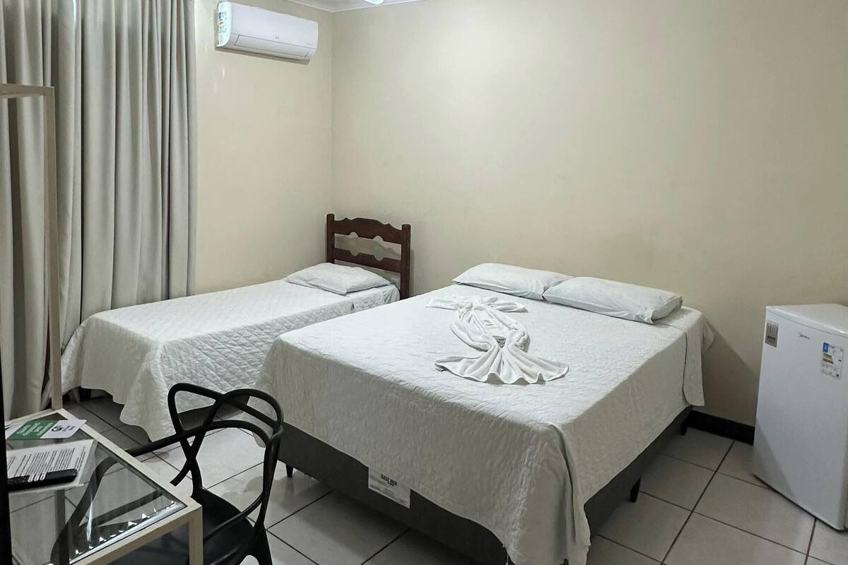 A hotel room with two beds, a ceiling fan, an air conditioning unit, a desk with a chair, and a refrigerator.