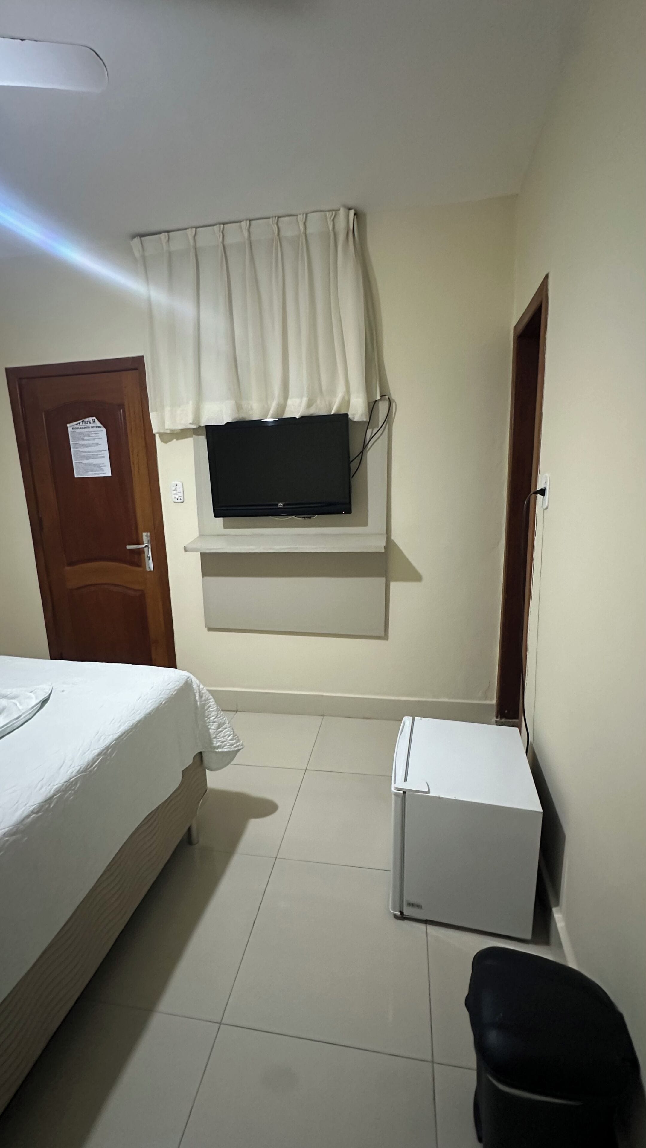 A bedroom with a bed, a television mounted on the wall, a small white cabinet, and a door.