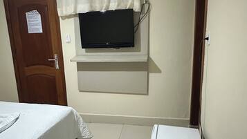 Quarto com uma cama, televisão na parede, um armário pequeno branco e uma porta.