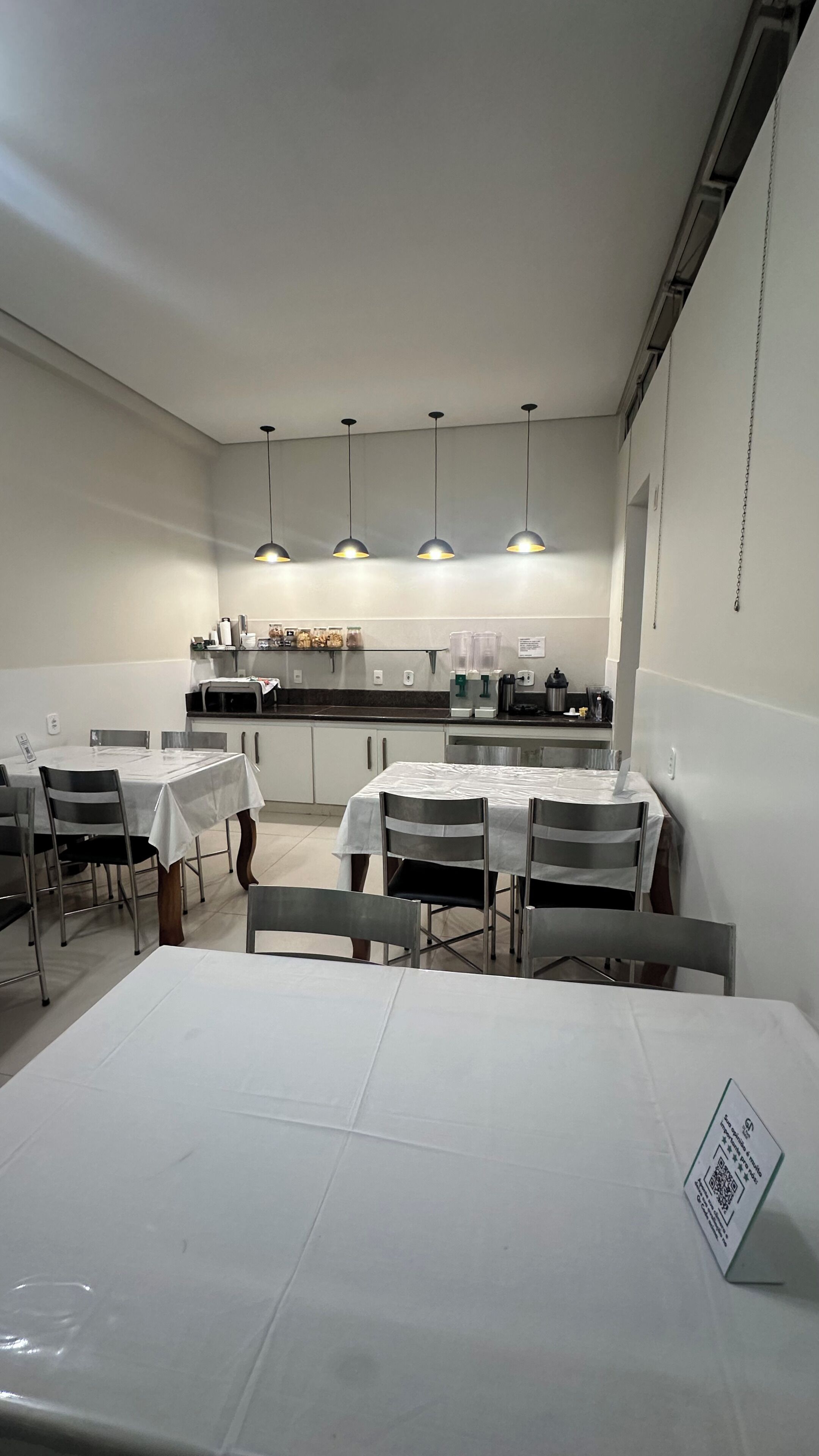 Zona pranzo con tavoli e sedie, cucina sullo sfondo e lampade a sospensione.