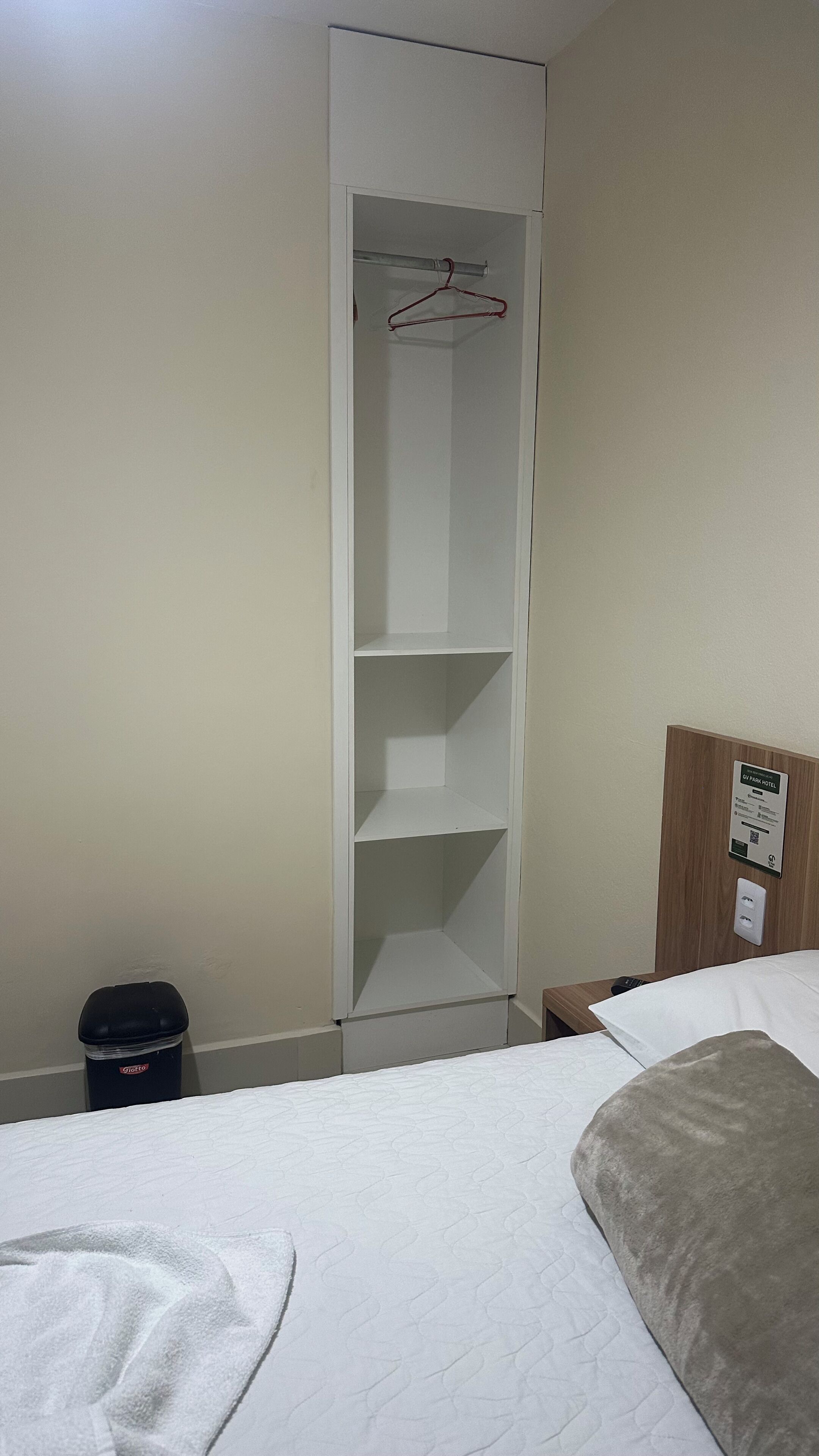 Quarto de hotel com cama, guarda-roupa, lixeira e cabeceira de madeira.