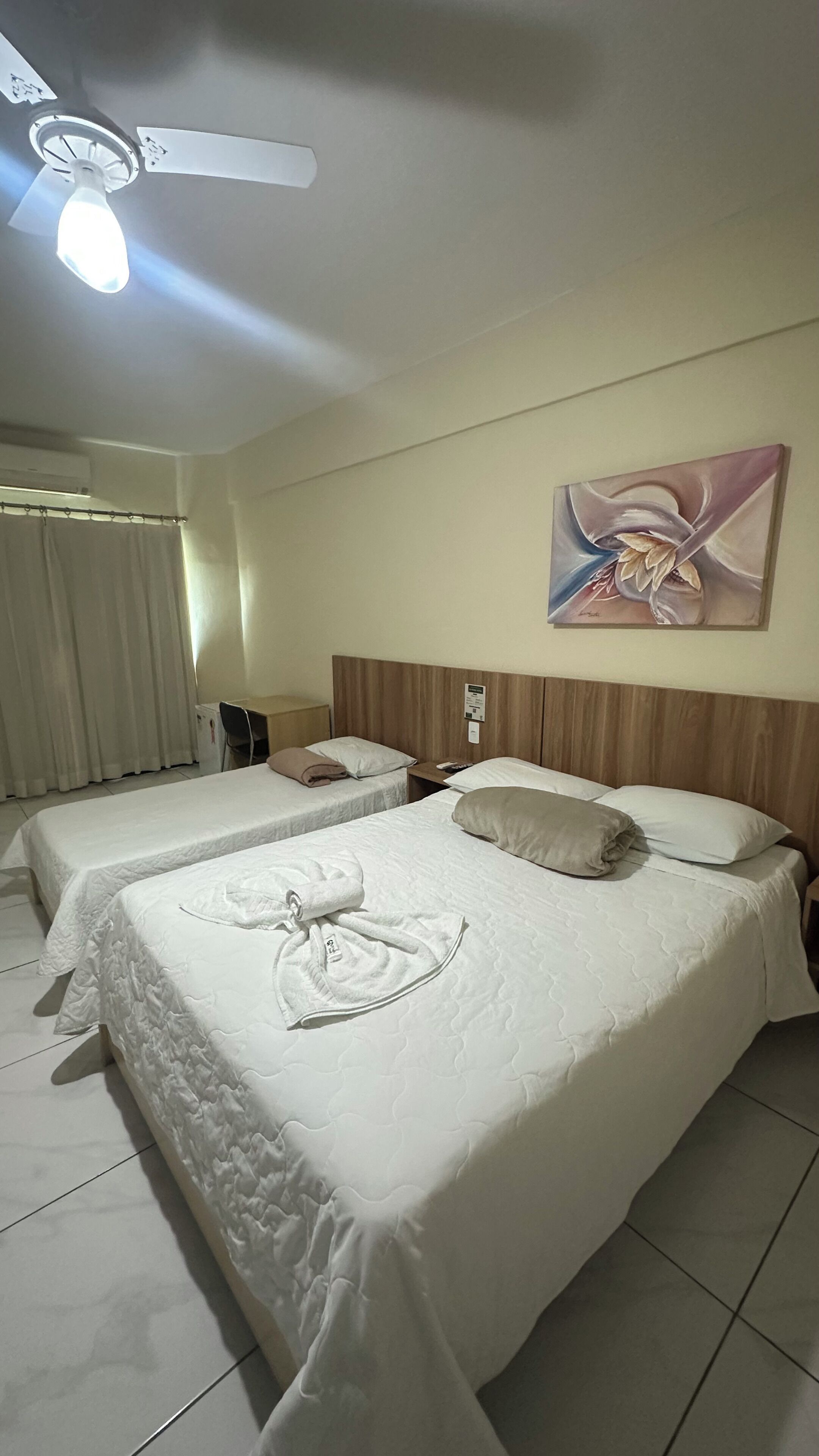 A hotel room with two beds, a ceiling fan, a painting, and a desk.