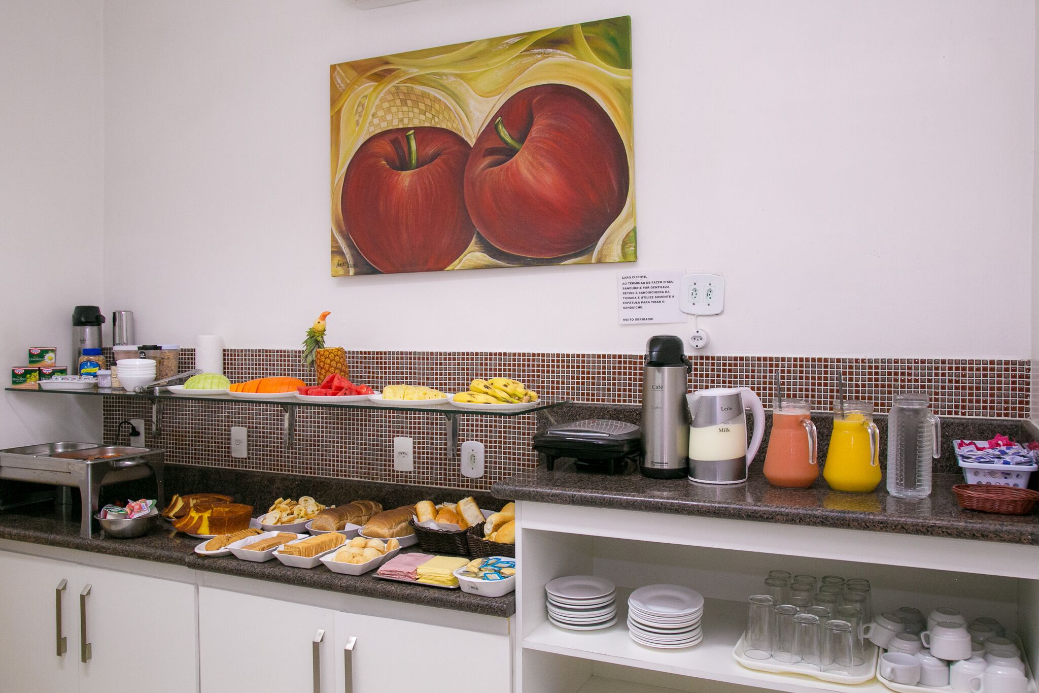 A breakfast buffet with various food items, a coffee maker, and a painting of apples on the wall.