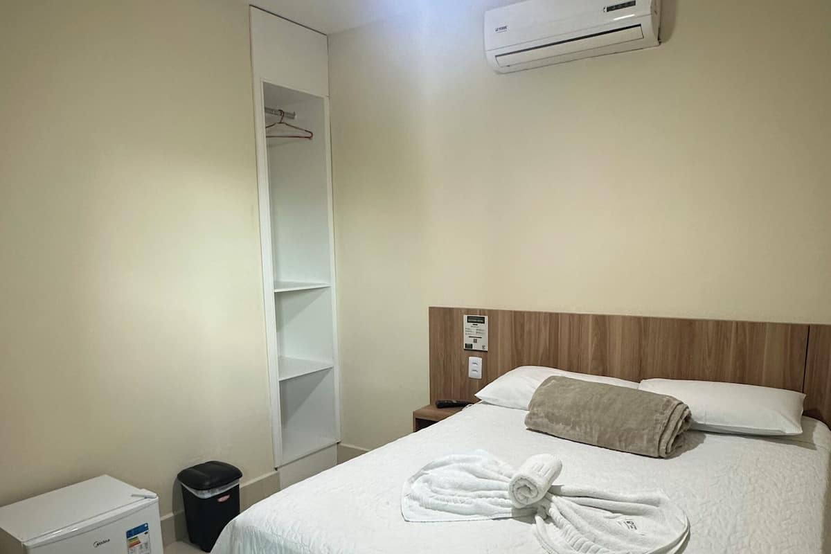 A hotel room with a bed, a ceiling fan, an air conditioning unit, a small table, and a trash bin.