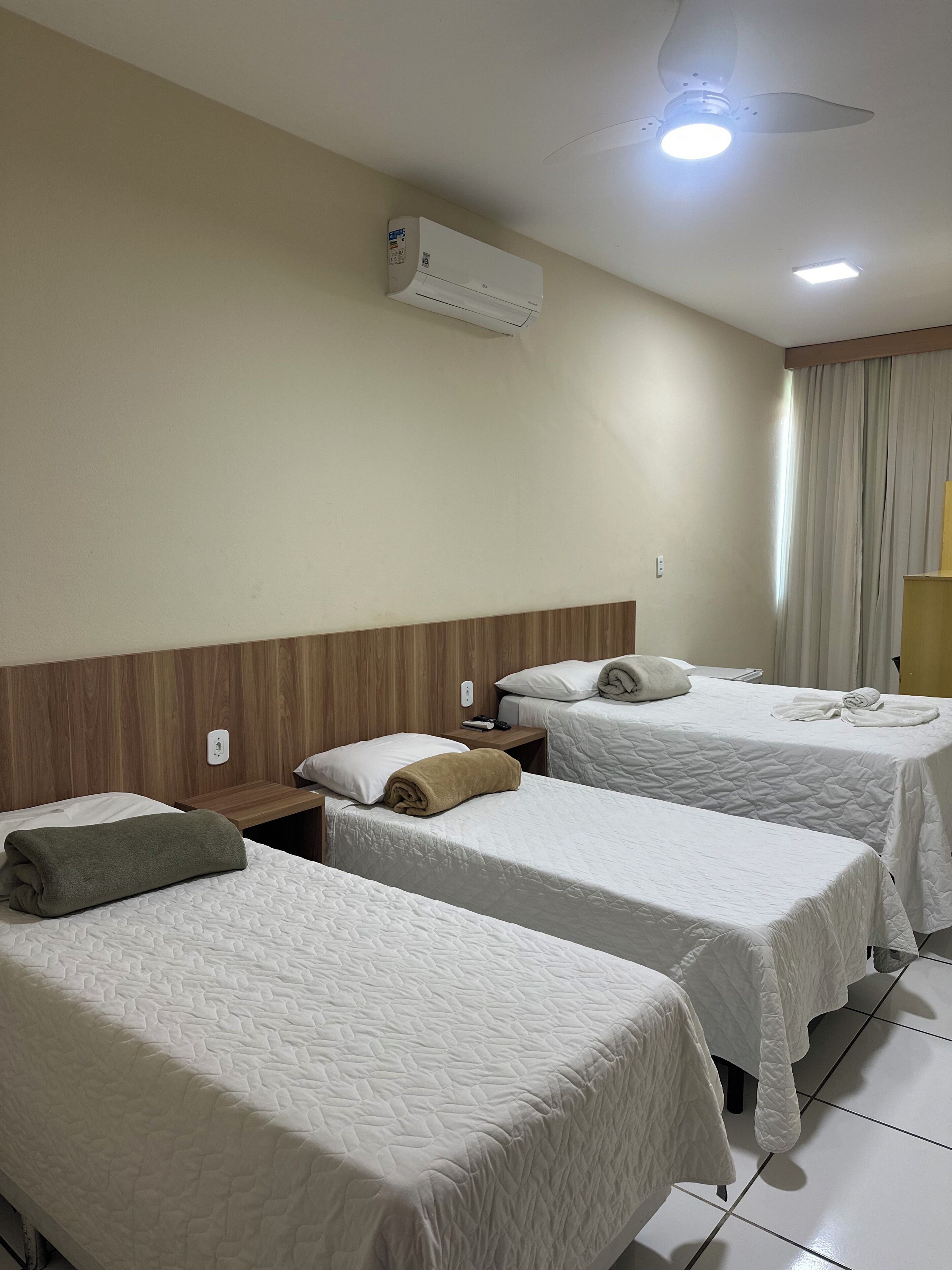 Habitación de hotel con dos camas individuales, ventilador de techo, unidad de aire acondicionado, mesita de noche con lámpara y ventana con cortinas.
