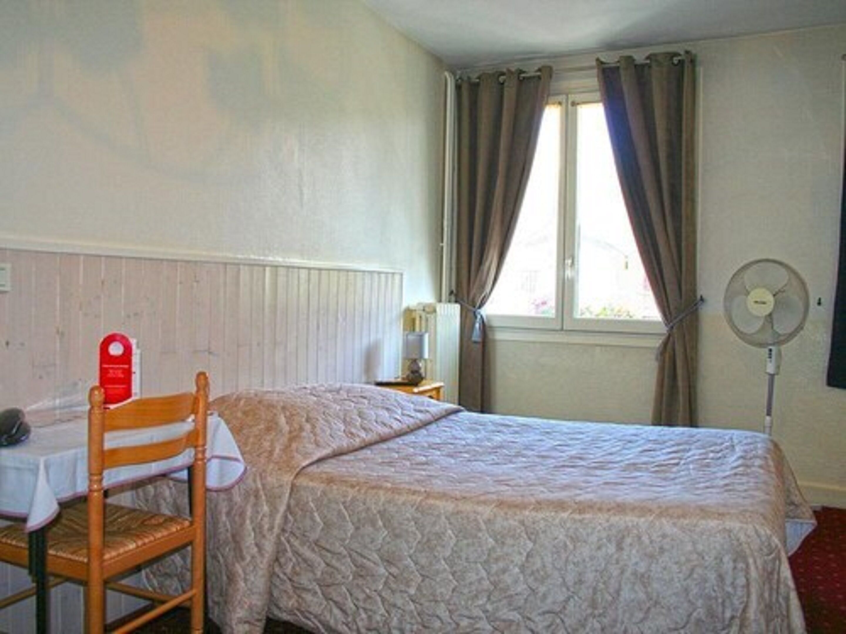 Quarto com cama, poltrona, mesa, abajur, janela com cortinas e ventilador fixado na parede.