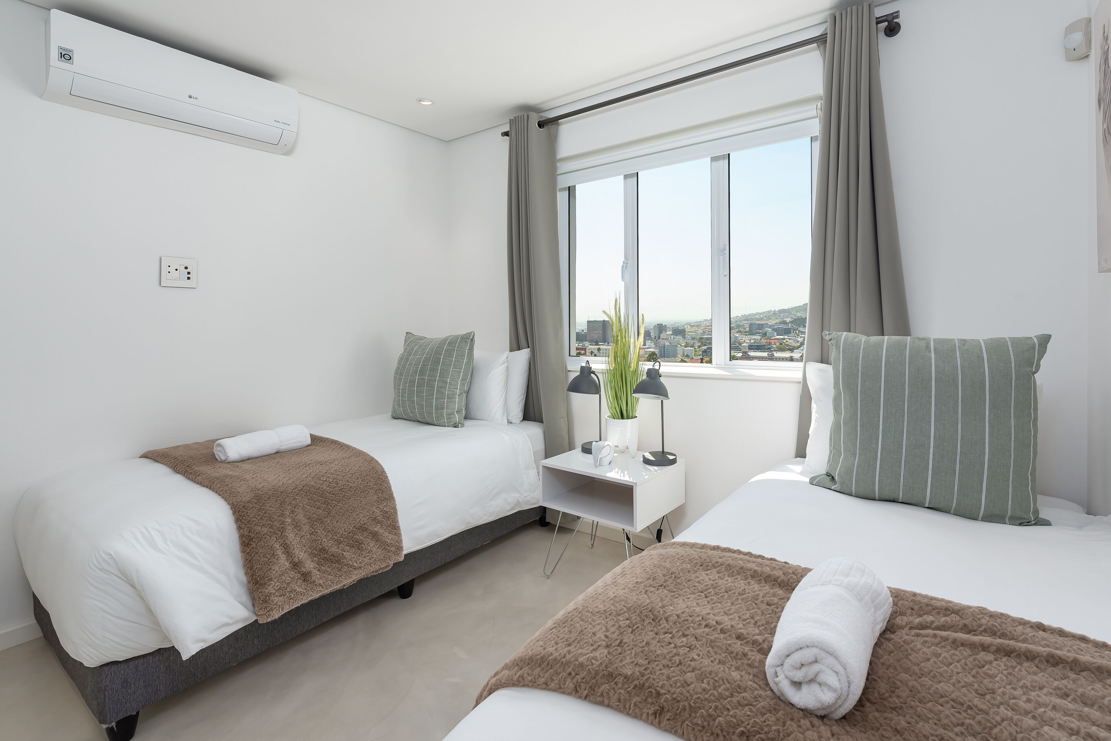 A modern hotel room with two beds, a bedside table, a window with a city view, and an air conditioning unit.