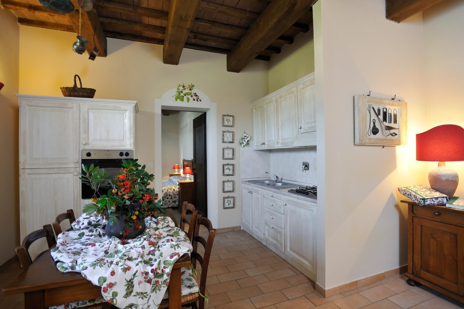 Una cocina con armarios blancos, una mesa de comedor con mantel floral y una lámpara roja sobre una mesita de madera.