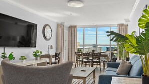 A modern living room with a flat-screen TV, a sofa, a dining table, and chairs. There is a large window with a view of the sea. - Brand New Premium Ocean View 3 Bedroom Condo w/ On-Site Rental Privileges (Myrtle Beach)