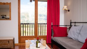 Ein Zimmer mit einem Bett, einem Tisch mit Weingläsern und einer Flasche, einem roten Vorhang und Blick auf einen Balkon mit Holzgeländer.