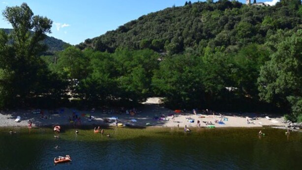A lakeside beach with people swimming and sunbathing, surrounded by dense forest and hills. - Camping La Charderie (Pont-de-Labeaume)
