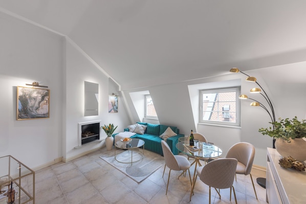 A modern living room with a teal sofa, a glass coffee table, and a dining area with chairs. There is a fireplace, a large window, and a ceiling light fixture. - President Boutique Apartments #8 by goodnite cz (Brno)