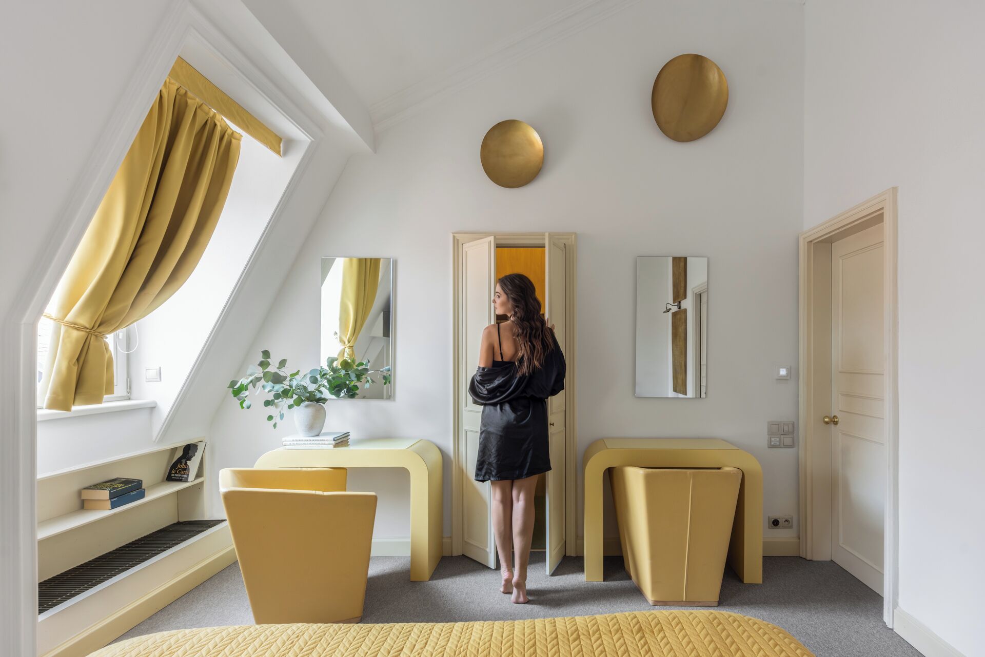 A modern bedroom with a sloped ceiling, a large window with yellow curtains, a bedside table with a potted plant, a mirror, and a door leading to another room.