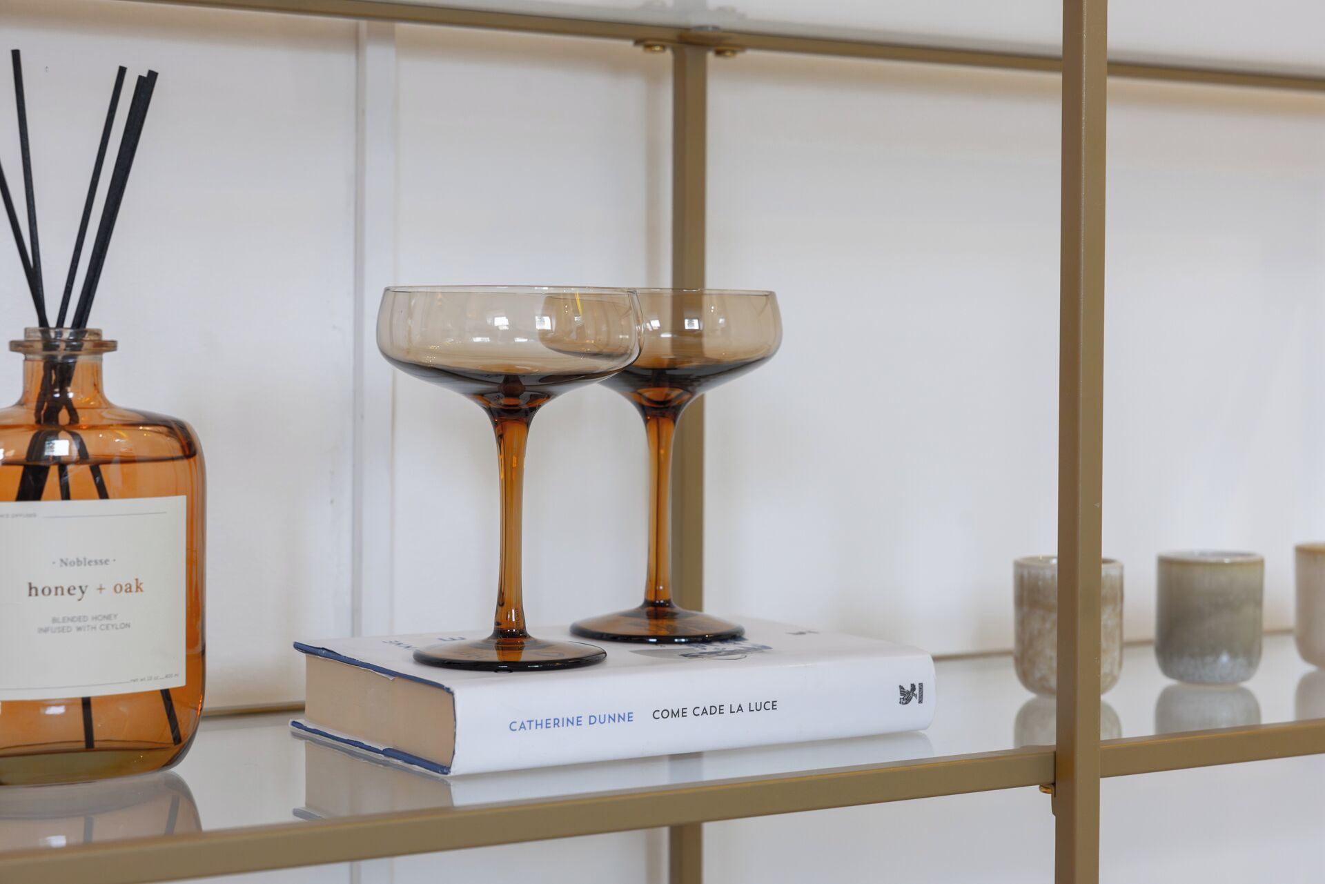 A glass shelf with a bottle of honey + oak, two wine glasses, and a book titled 'Come Cade La Luce' by Catherine Dunne.