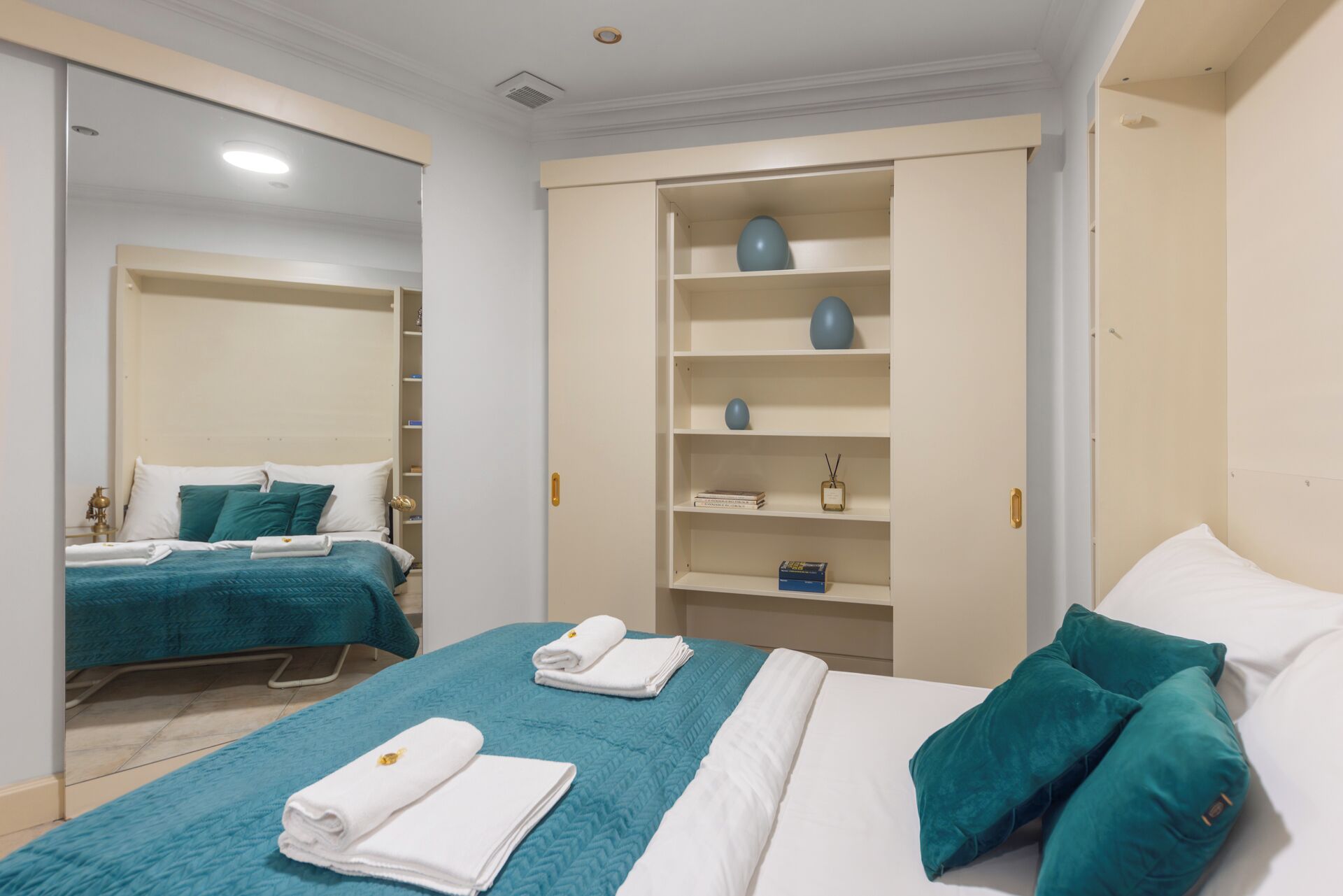 A hotel room with two beds, teal bedding, white pillows, a wardrobe with shelves, and a mirror.