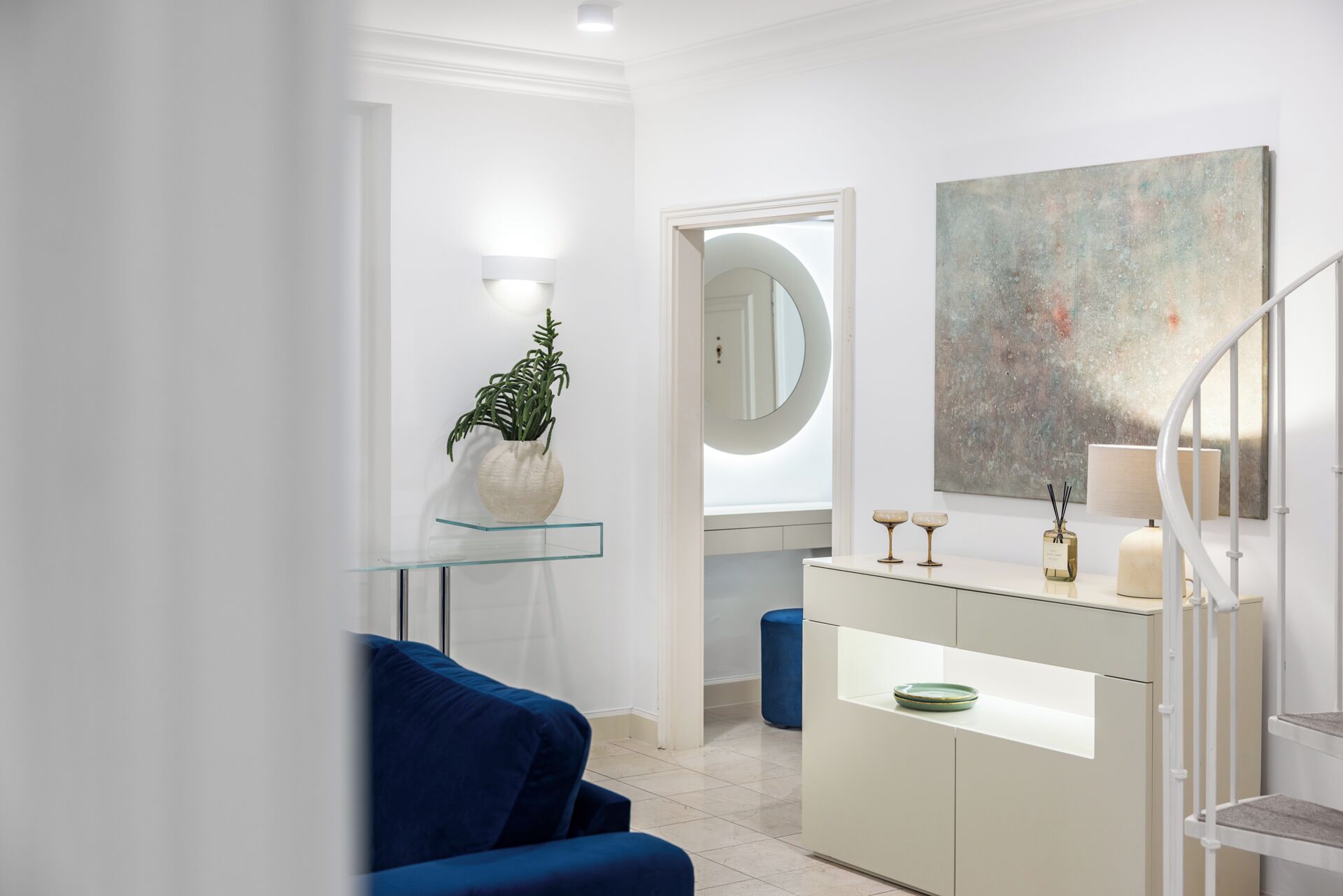 Et moderne badeværelse med en glashylde, en blå stol, et hvidt toiletbord og et stort abstrakt maleri.