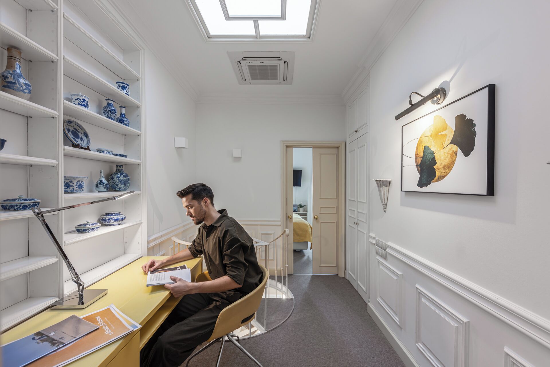 A man sitting at a desk in a room with white walls, a skylight, and shelves with blue and white pottery.