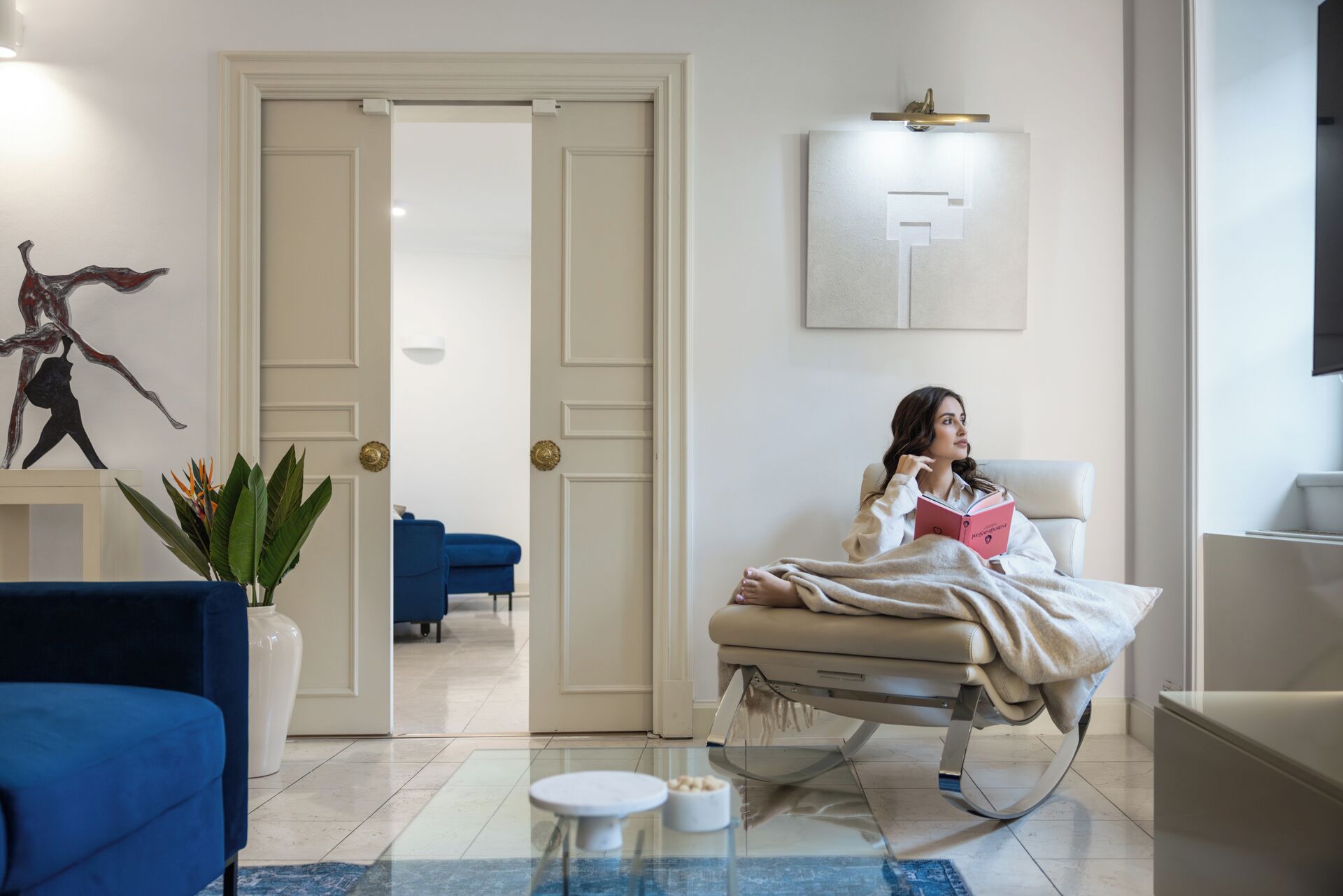 A modern room with a bed, a blue armchair, a white bedside table, and a plant.