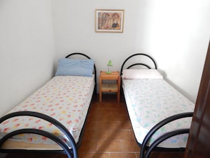 A small room with two single beds, each with a floral bedspread, a nightstand with a lamp, and a painting on the wall. - Mono Istana (Olbia)