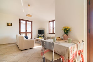 A living room with a white sofa, a dining table with a floral tablecloth, and a television on a stand.