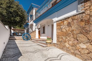 A narrow cobblestone street with a stone wall on one side and a white building with blue trim and a balcony on the other. - Casa Festa (Dorgali)