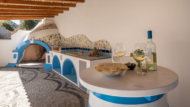 A cozy outdoor seating area with a mosaic-tiled wall, a wooden bench, and a table with a bottle of white wine and glasses.