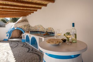 A cozy outdoor seating area with a mosaic-tiled wall, a wooden bench, and a table with a bottle of white wine and glasses. - Casa Festa (Dorgali)
