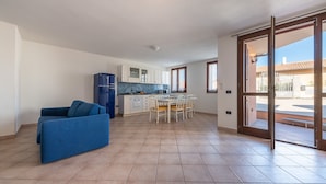 A spacious living area with a blue sofa, a dining table with chairs, a kitchen with white cabinets, and a sliding glass door leading to an outdoor area. - Casa Tortora (Siniscola)