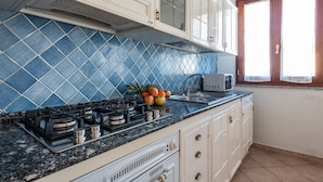 A kitchen with a gas stove, a microwave, and a sink. There are cabinets with glass doors and a window with curtains. - Casa Tortora (Siniscola)