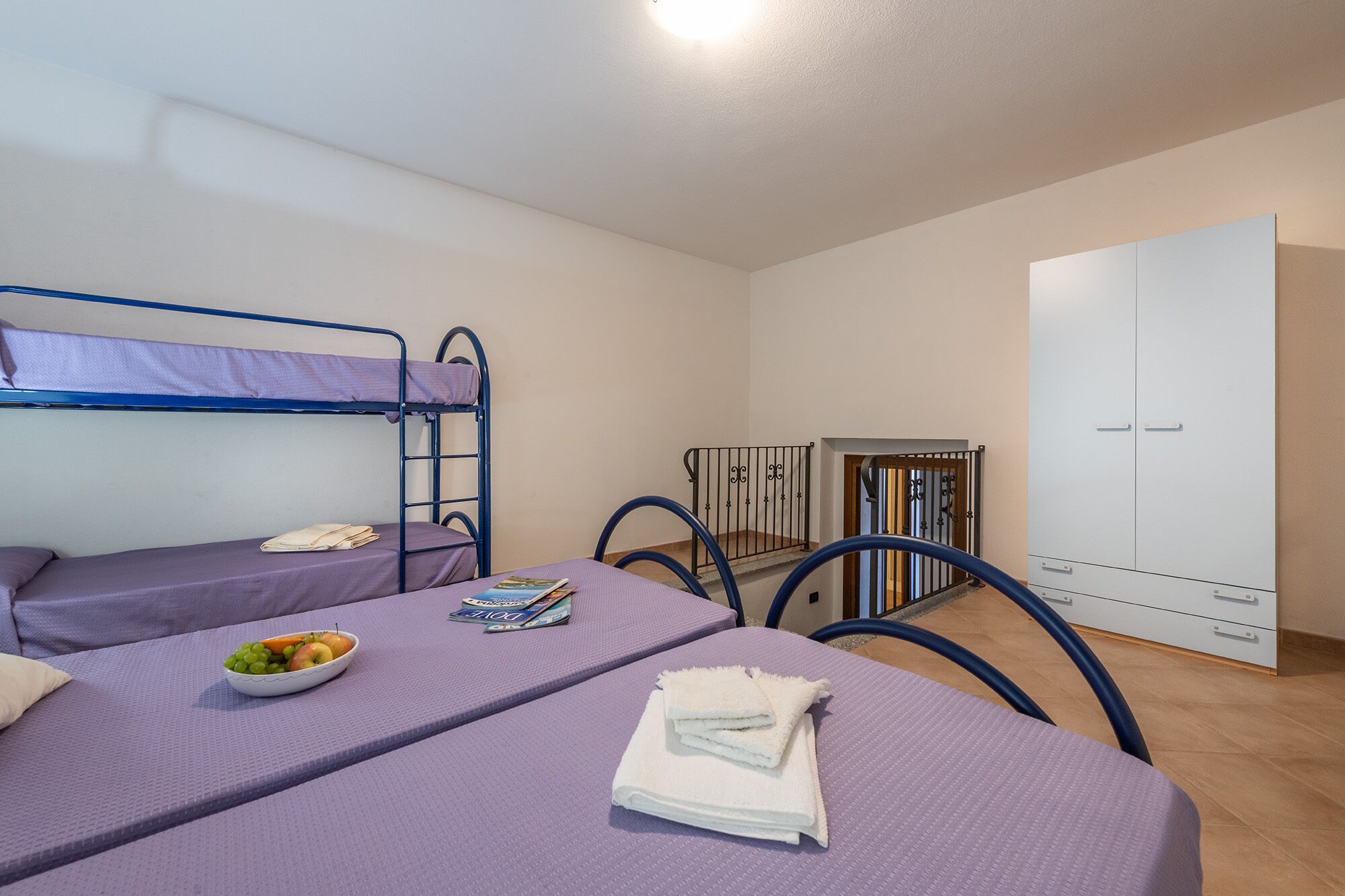 A dormitory room with bunk beds, a desk with a chair, a wardrobe, and a small table with a bowl of fruit and some books.