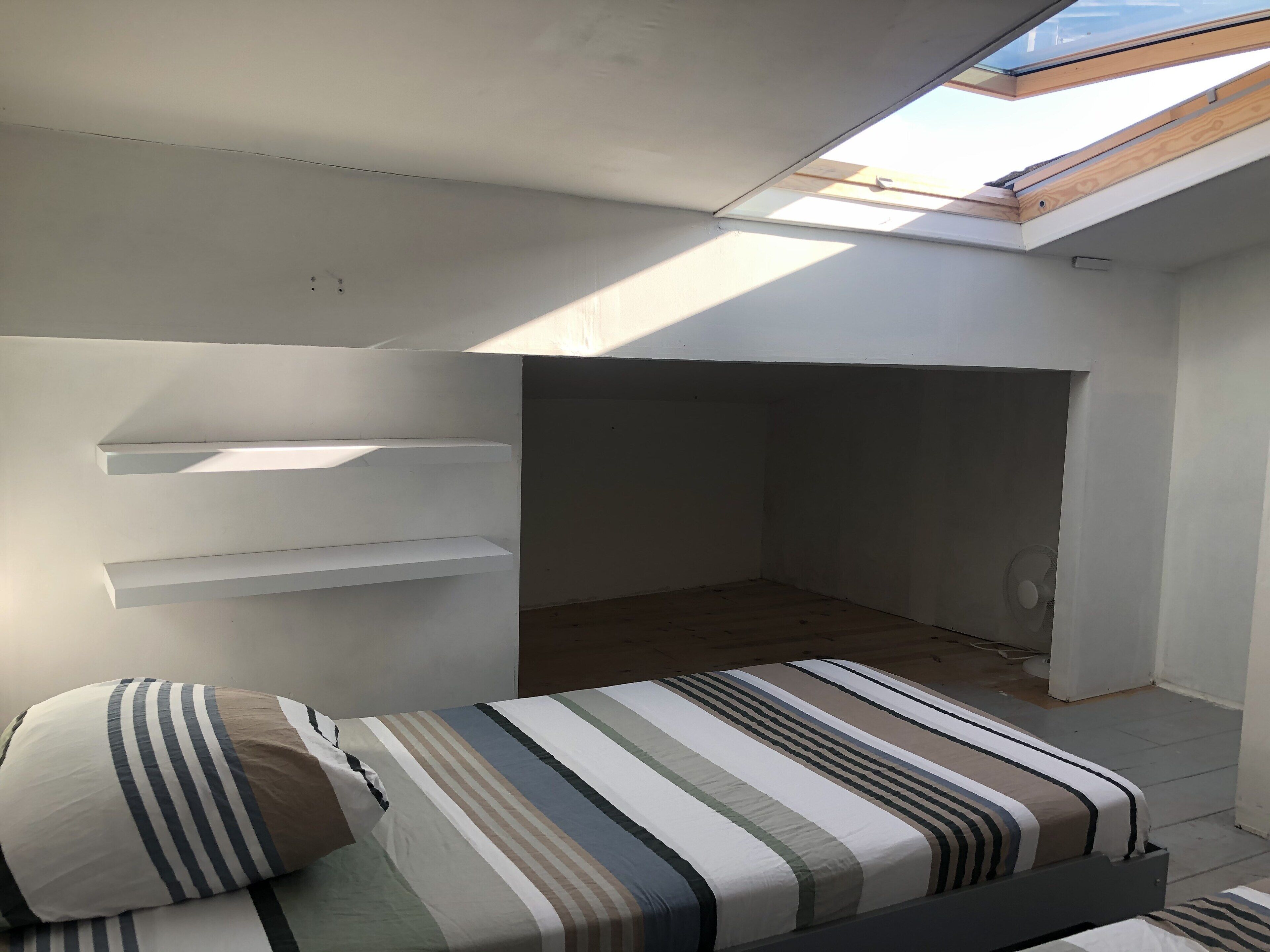 Een kleine, lichte kamer met dakraam, een bed met gestreept beddengoed en een ventilator.