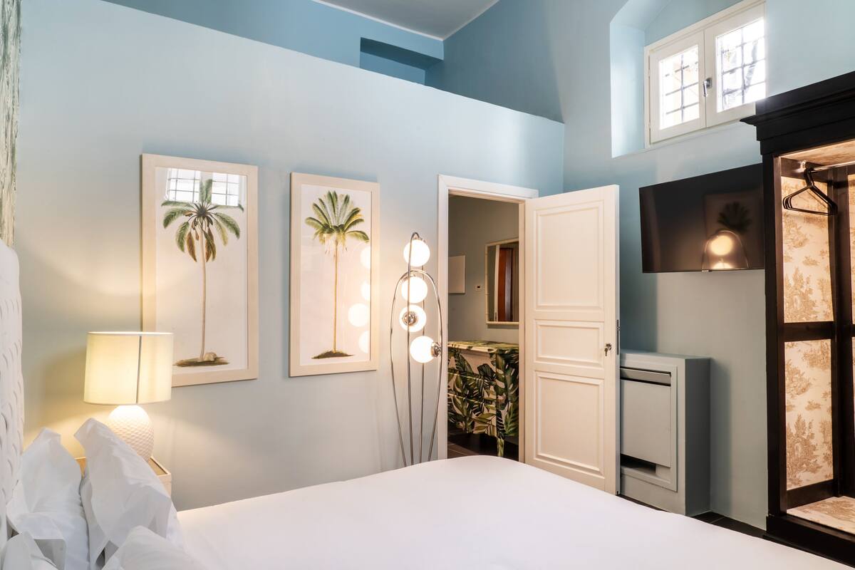 A bedroom with a bed, a nightstand with a lamp, framed artwork of palm trees, a mirror, and a door leading to a bathroom.