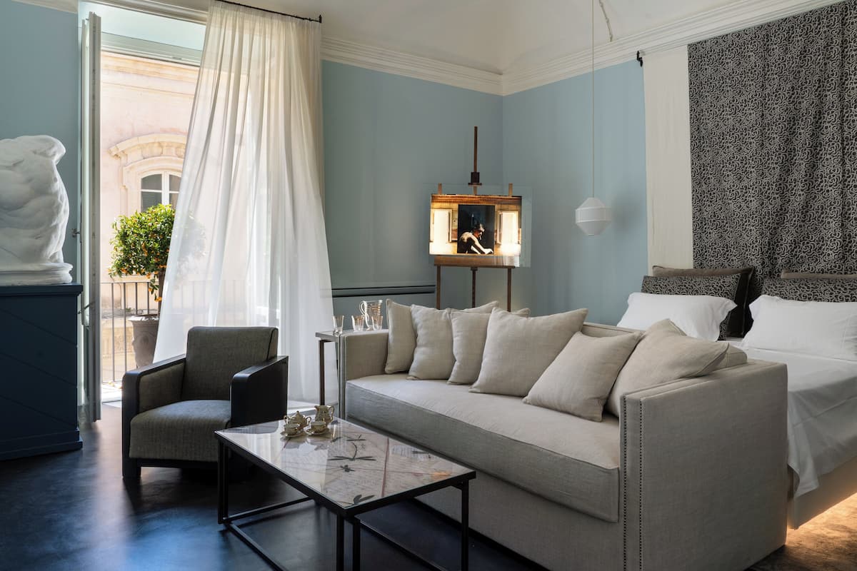 A modern hotel room with a sofa, armchair, and a small table. There is a large window with sheer curtains, a potted plant, and a view of a building outside.