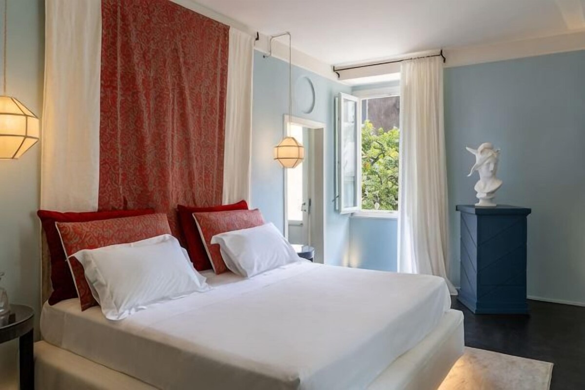 A bedroom with a large bed, two bedside lamps, a window with curtains, and a statue on a pedestal.