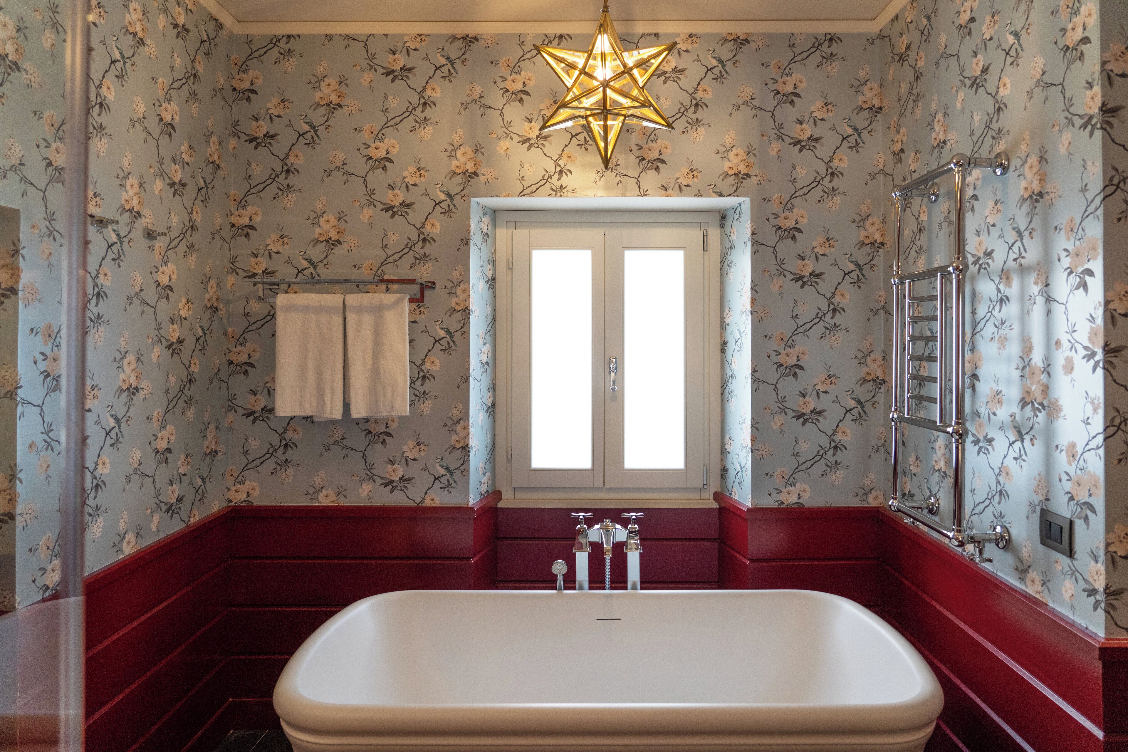 A bathroom with floral wallpaper, a freestanding bathtub, a star-shaped light fixture, and a window with a towel rack.