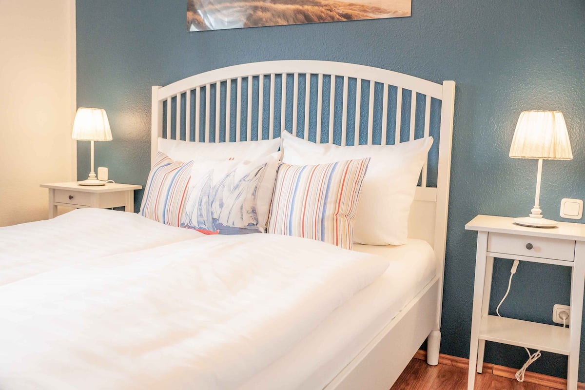 A neatly made bed with white bedding and striped pillows, two bedside tables with lamps, and a blue wall.