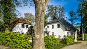 A white house with a sloped roof and a balcony, surrounded by greenery and a dirt path.