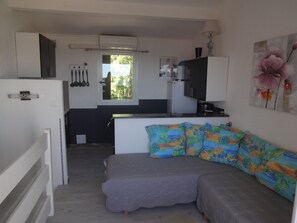 Ett kompakt bostadsutrymme med ett litet kök, en grå soffa med färgglada kuddar och ett fönster med utsikt över grönområden.