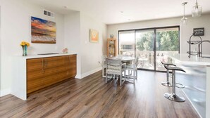 A modern kitchen with a wooden cabinet, a dining table with chairs, and a large window offering a view of greenery.