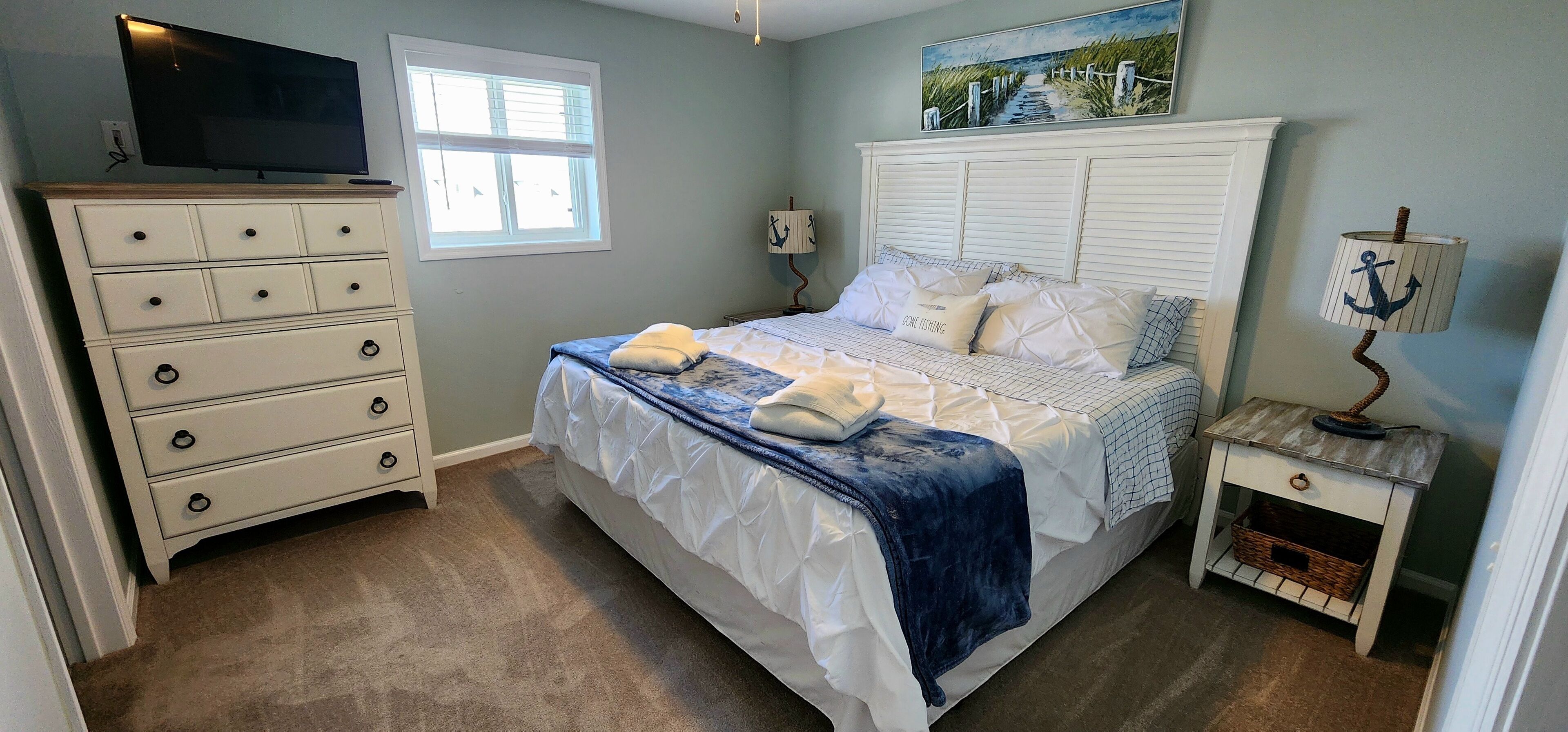 A bedroom with a bed, dresser, nightstand, lamp, and a painting on the wall.