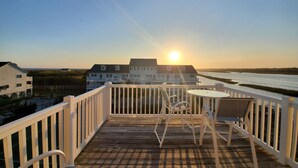 A wooden deck with white railings, a table, and two chairs. In the background, there is a white building with a balcony, and a river with a bridge is visible. - Spectacular Sunset Views  from this Top/3rd floor condo, 2 bed/2 bath (Ocean Isle Beach)
