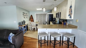 A modern kitchen with a central island, bar stools, and a dining area with a chair and table. - Spectacular Sunset Views  from this Top/3rd floor condo, 2 bed/2 bath (Ocean Isle Beach)