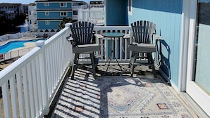 A balcony with two Adirondack chairs and a small table, overlooking a swimming pool and adjacent buildings. - Spectacular Sunset Views  from this Top/3rd floor condo, 2 bed/2 bath (Ocean Isle Beach)