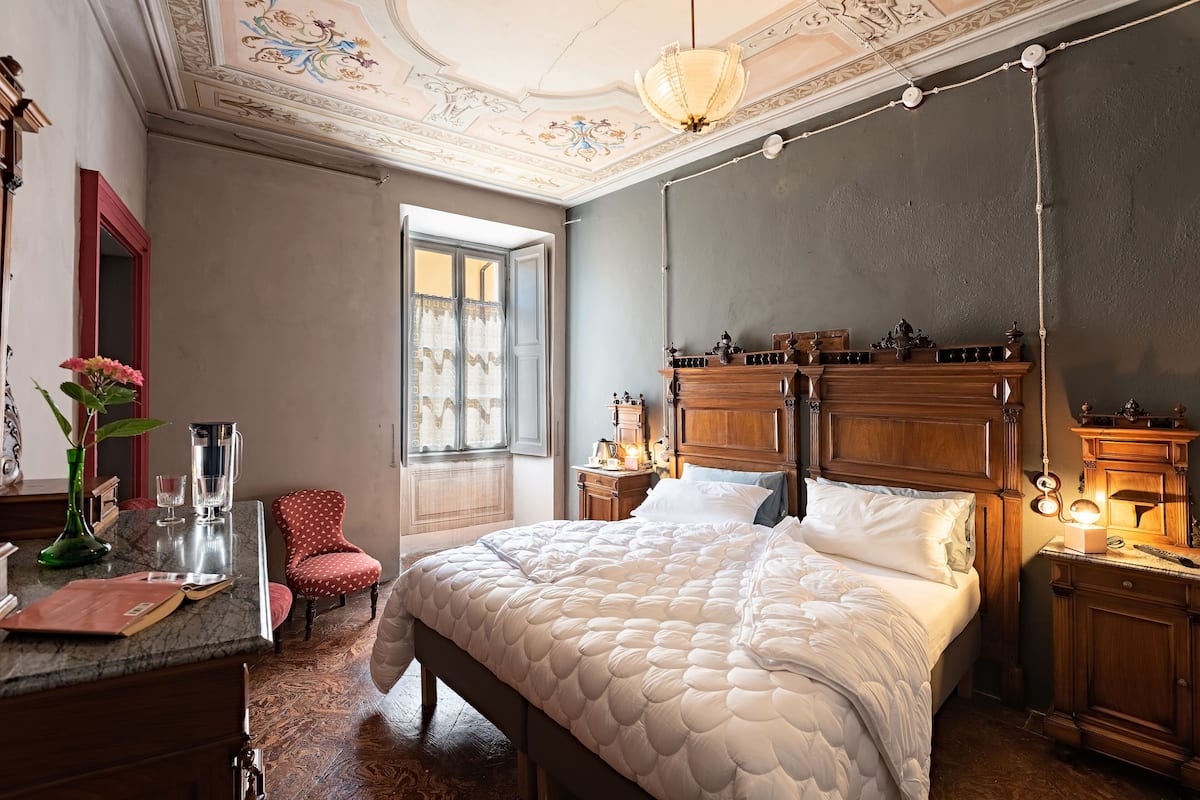 A bedroom with a large bed, a wooden nightstand, a desk with a vase, and a chair.