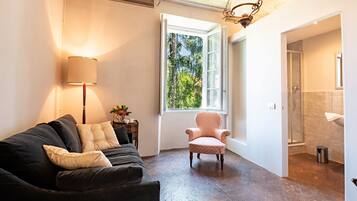Un salon avec un canapé noir, un fauteuil rose, une lampe et une fenêtre donnant sur des arbres.