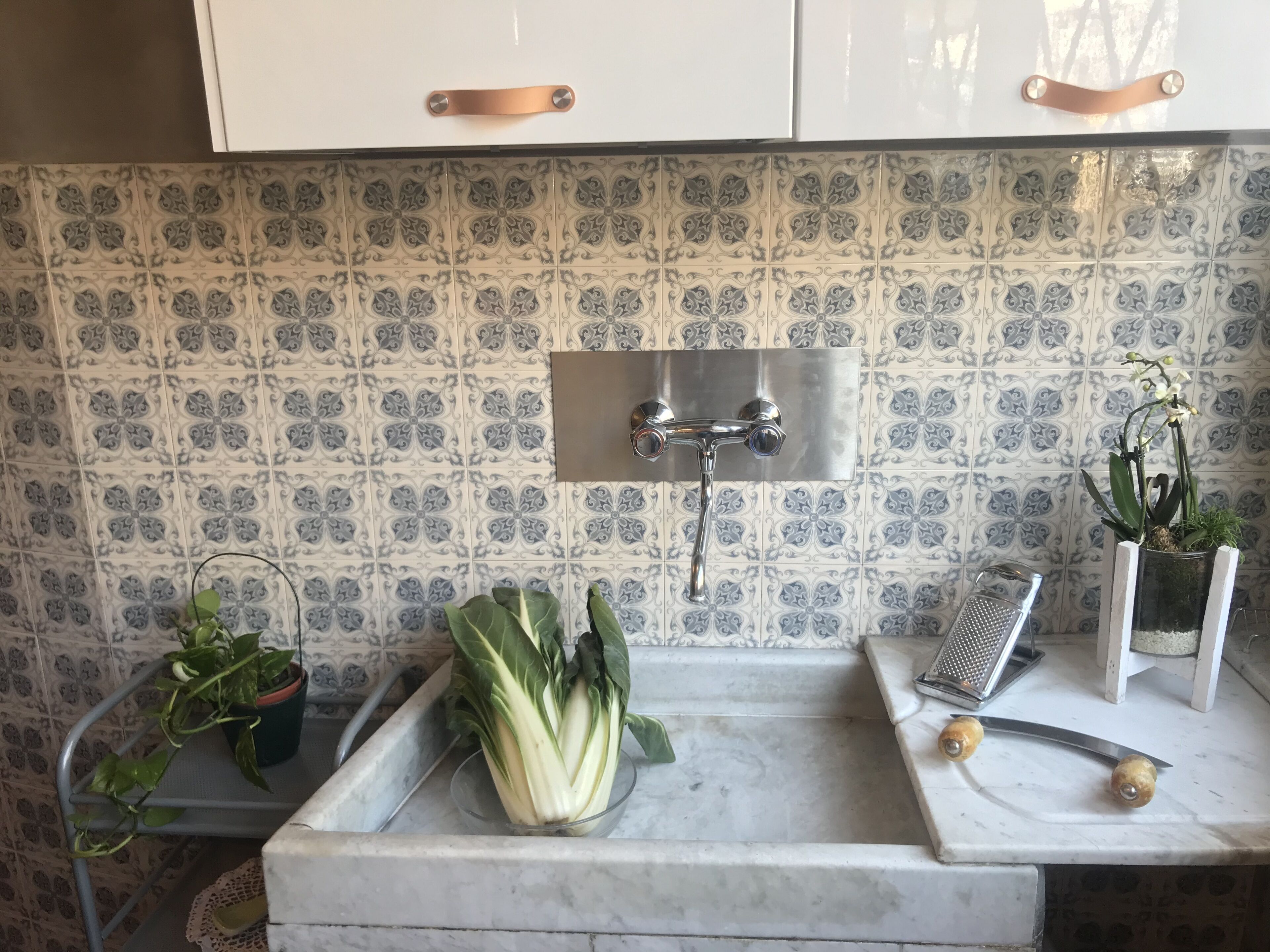 A kitchen with a sink, a potted plant, a bowl of vegetables, and a grater on the counter.