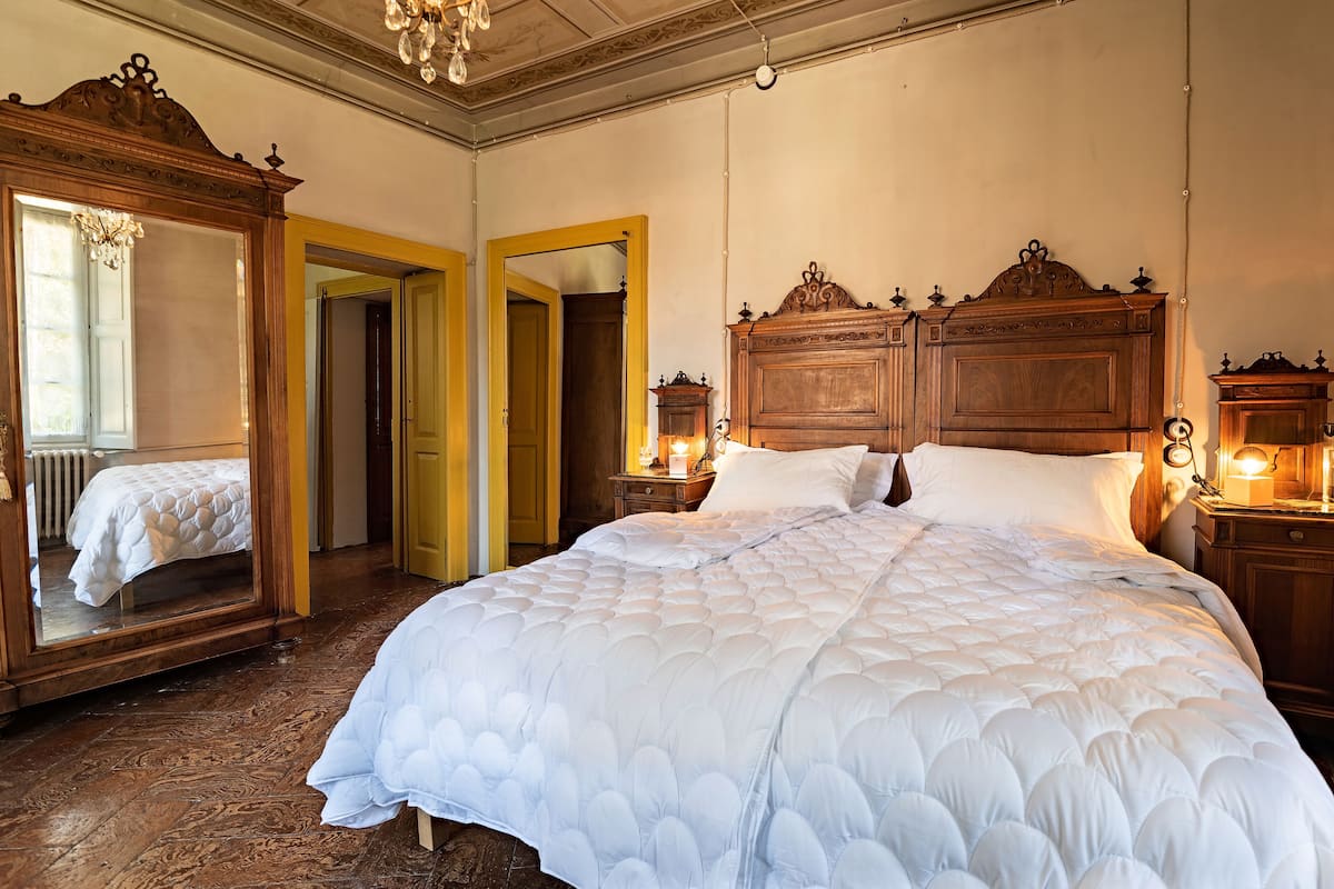 A large bed with a white quilt, wooden headboard, and bedside lamps. There is a mirror and a door leading to another room.