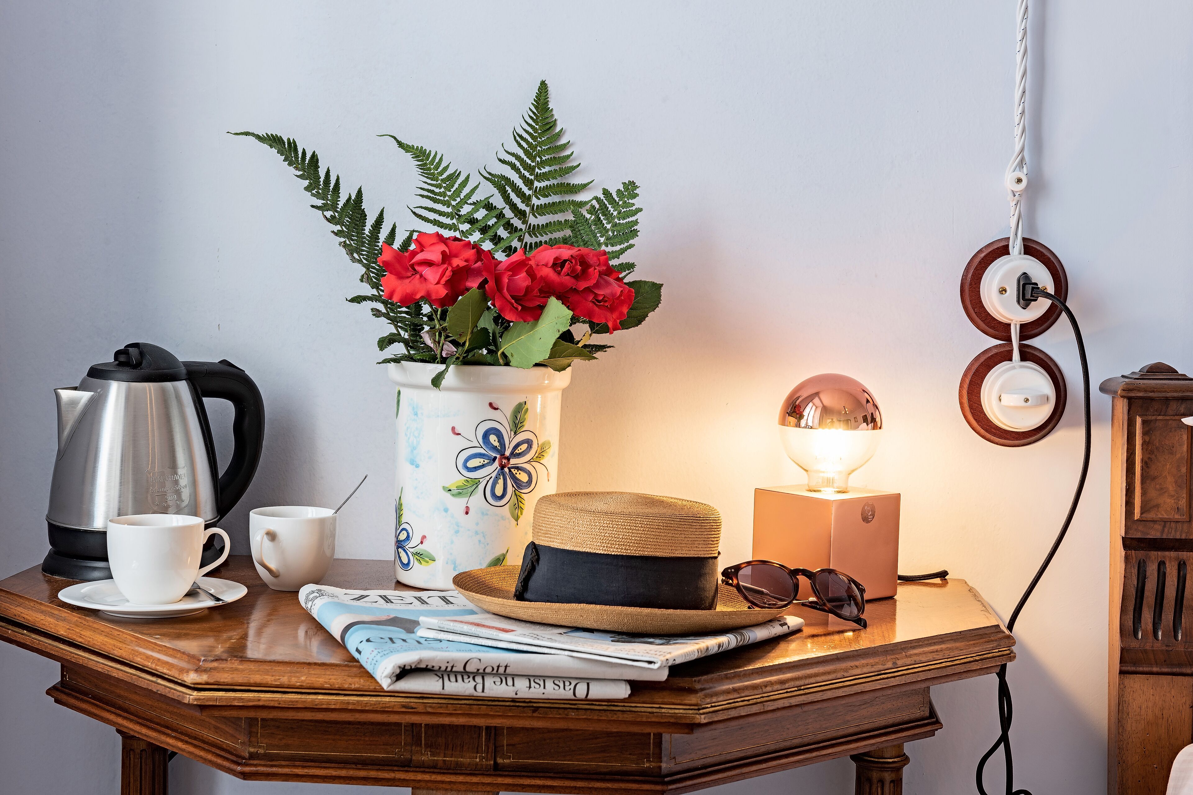 A wooden table with a coffee pot, cups, a hat, sunglasses, a lamp, a flower vase, and a magazine.