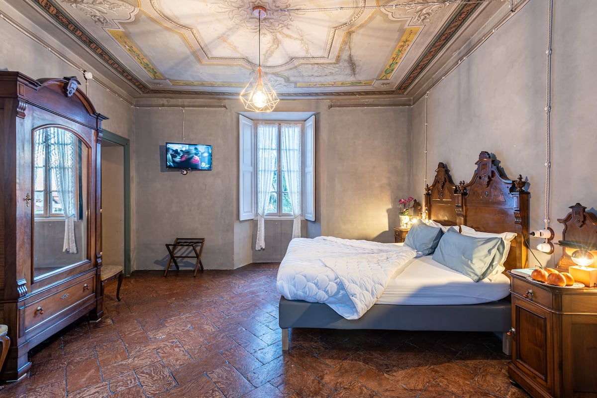 A bedroom with a large bed, a wooden wardrobe, a television on the wall, and a window with curtains.