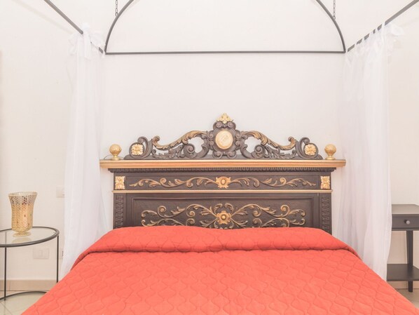 Un dormitorio con una cama, un cabecero decorativo, una pequeña mesa redonda con un florero y una cortina.